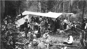 Men and women sitting, standing, and eating in and around a canvas tarp in the woods